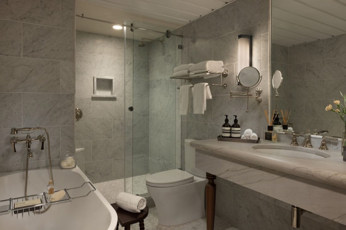 The bathroom in Room Five, a roll-top tub in marble beside a glass shower and a shaving mirror.